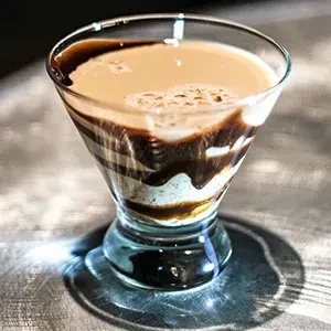 Clear martini glass with layered chocolate, cream, and frothy coffee beverage.
