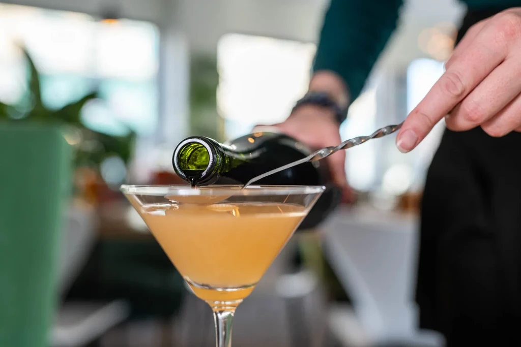 Hands pour liquid into an amber cocktail in a martini glass.
