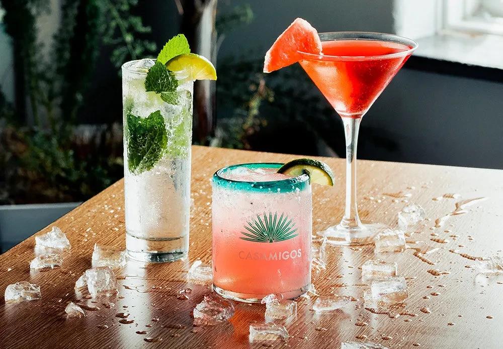 Three cocktails with Casamigos logo, mint, and watermelon on wooden table.