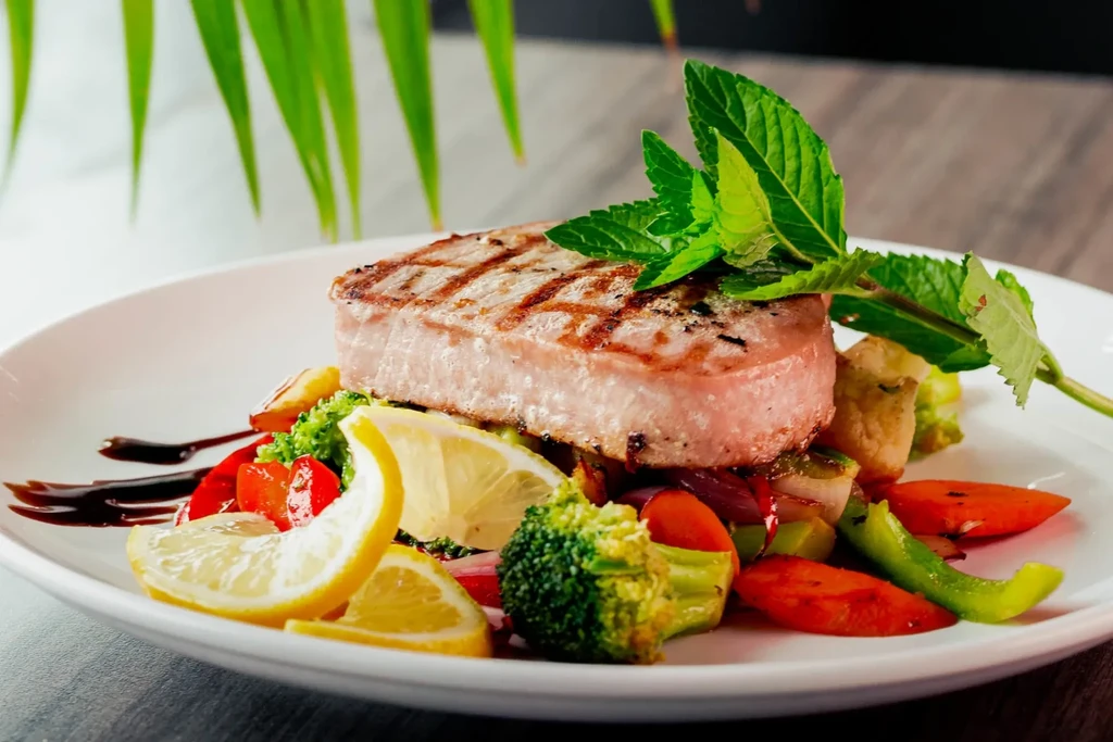 Grilled meat, colorful vegetables, mint leaves, and lemon wedges on white plate.