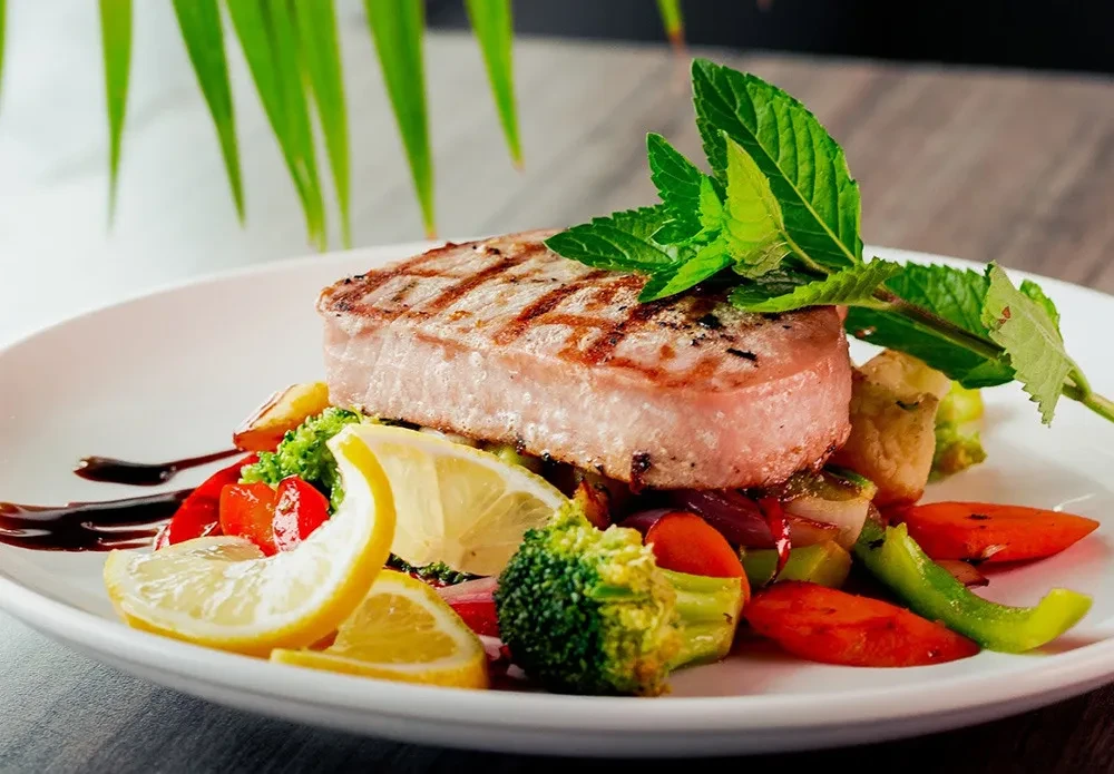 Grilled tuna steak, colorful vegetables, lemon, mint, and dark sauce.