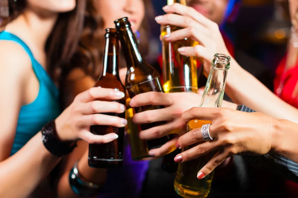 Close-up of hands toasting with beer bottles in dim, colorful light.
