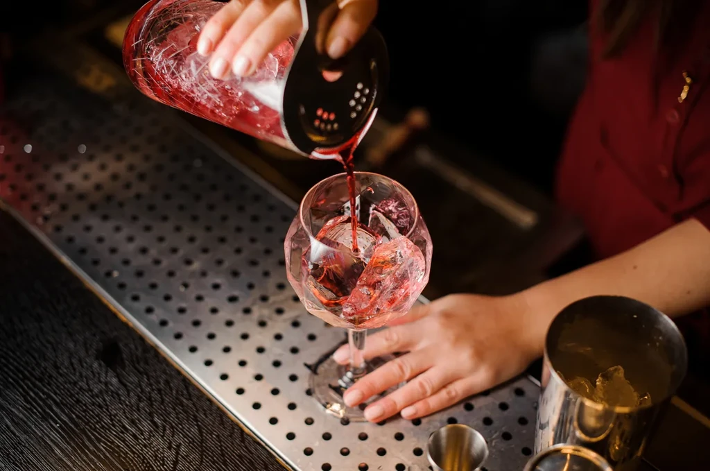 Bartender's hands pour vibrant red cocktail over ice into faceted glass.