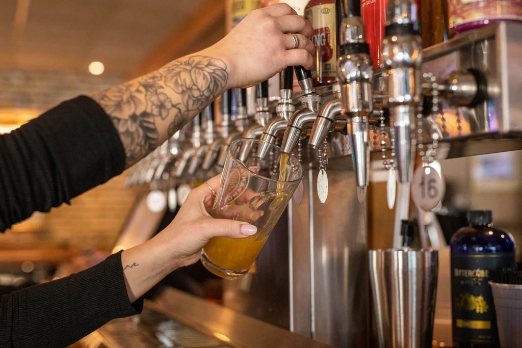 Hands with tattoos pouring beer/cider from bar tap into a glass.