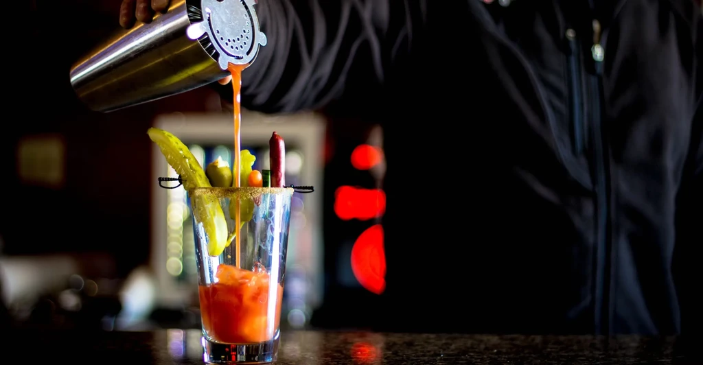 Bartender pouring reddish cocktail with pickle, olives, and sausage.