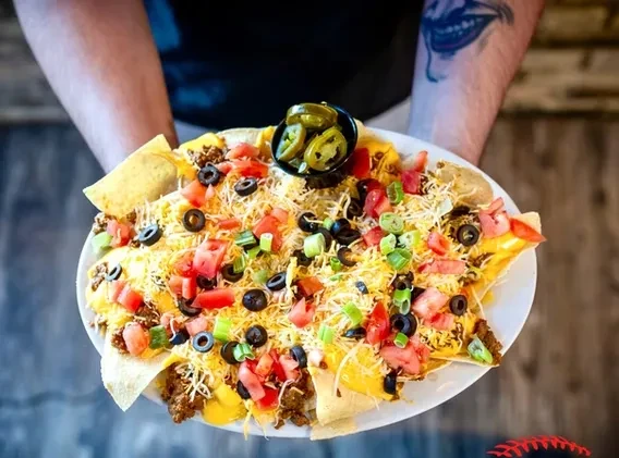 Plate of loaded nachos with jalapeños, held by arms, one tattooed.