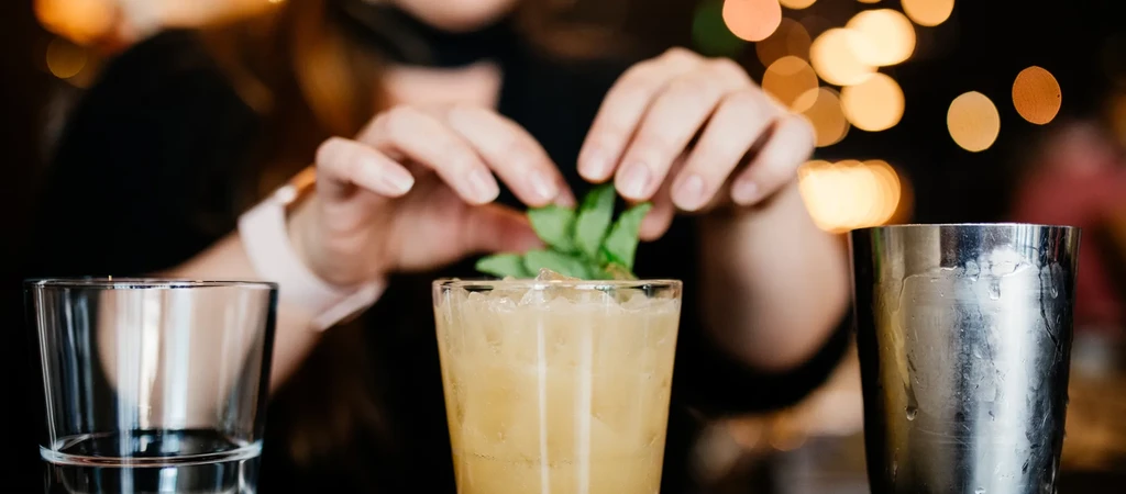 Close-up: hands garnish cocktail with fresh mint, shaker nearby.