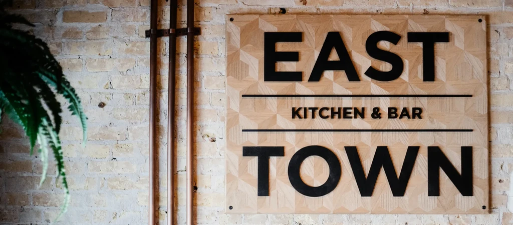Wooden sign "EAST KITCHEN & BAR TOWN" on brick wall, copper pipes.