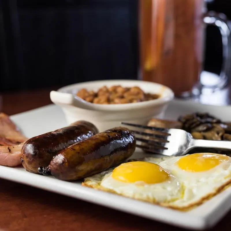 Rectangular white plate with sausages, fried eggs, bacon, mushrooms, and beans.