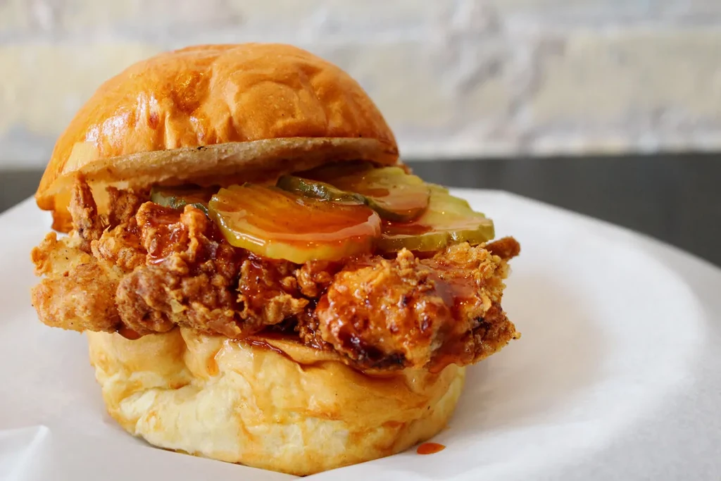 Crispy fried chicken sandwich with pickles and glossy reddish-orange sauce.