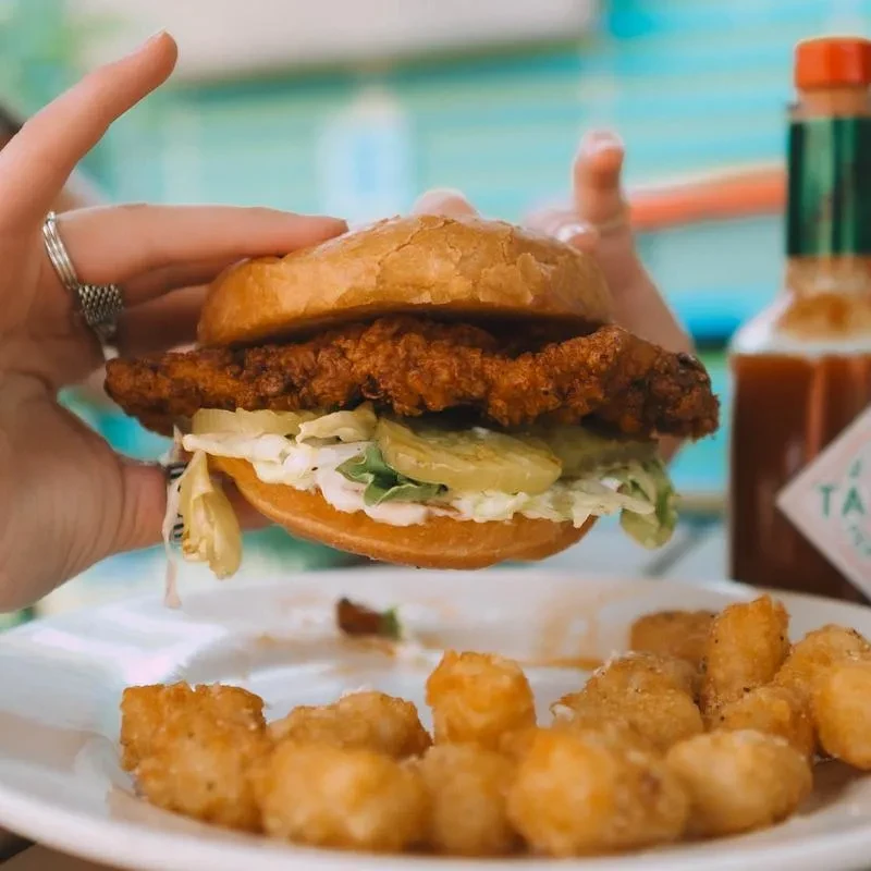Hands hold a crispy fried chicken sandwich with lettuce, pickles, and tater tots.