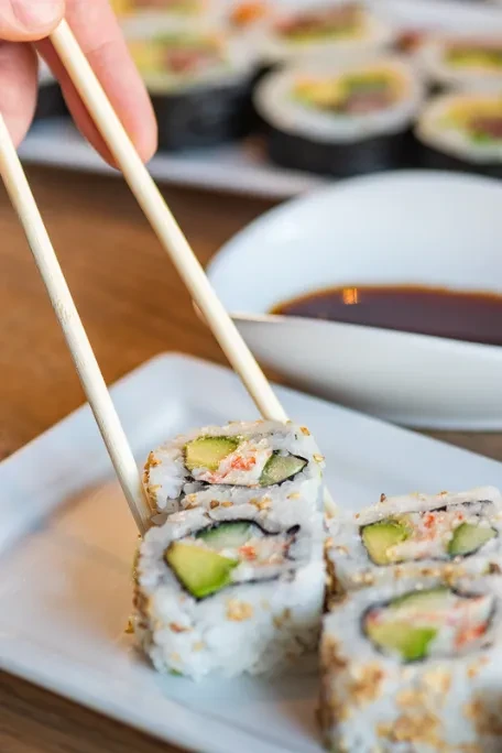 Hand picking a California sushi roll with chopsticks from a white plate.