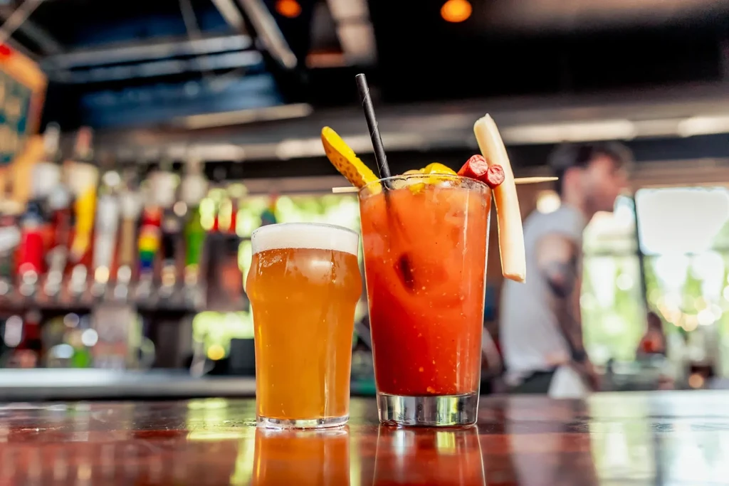 Amber beer and a Bloody Mary with garnishes on a wooden bar.