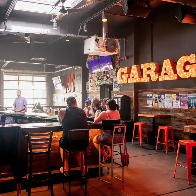Industrial-rustic bar with patrons, a bartender, and a large "GARAGE" sign.