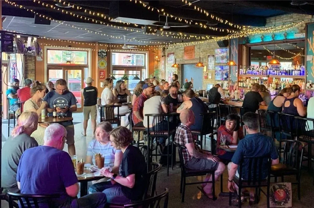 Bustling bar interior with patrons socializing, drinking, and a blue-lit bar.