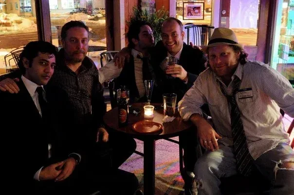 Five men at a bar, some smiling, one playfully kissing a cheek.