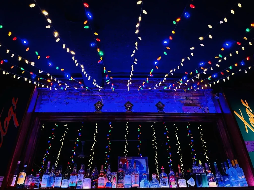 Bar interior featuring colorful string lights, liquor bottles, and a brick wall.