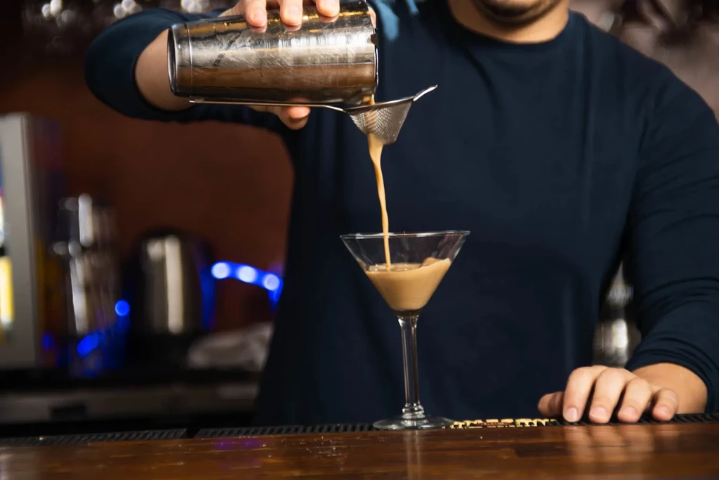 Bartender straining creamy brown cocktail into martini glass.