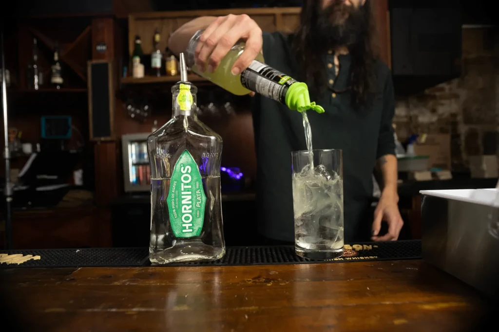 Hands pour drink into iced glass next to a Hornitos tequila bottle.