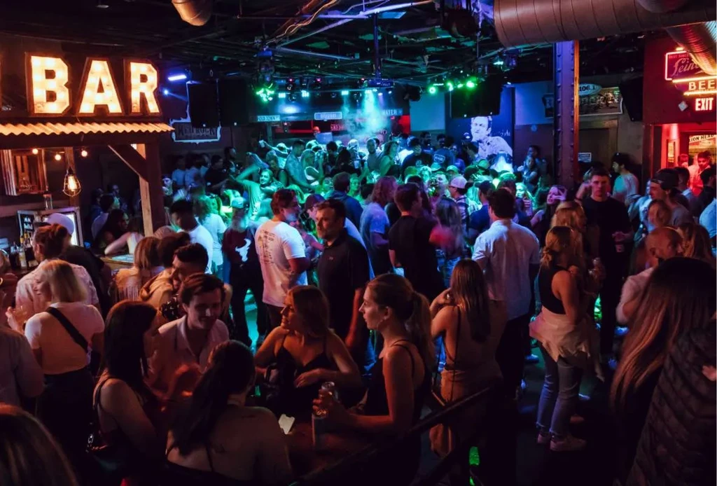 Lively, crowded bar with colorful neon lights and a BAR sign.