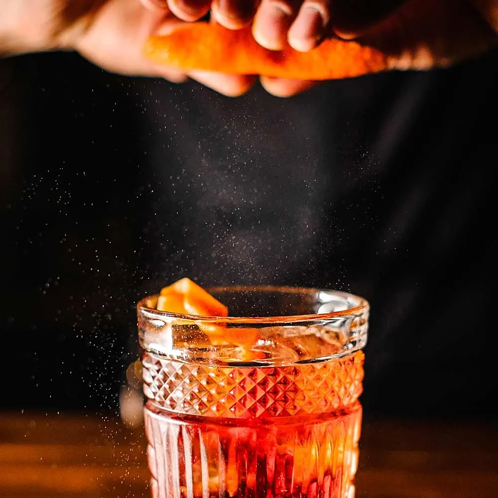 Orange cocktail garnished by hand with sparkling citrus oils spraying.