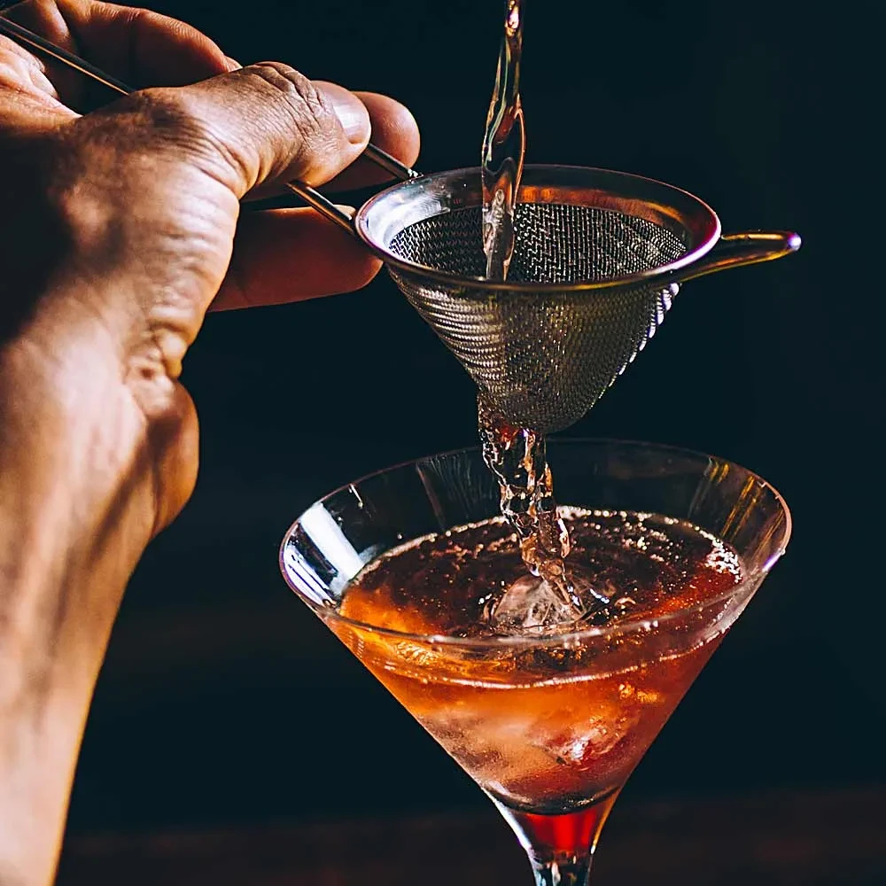 Bartender's hand straining amber drink into a martini glass with ice.