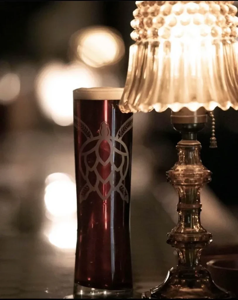 Dark beer glass with hop-turtle etching, by a glowing vintage lamp.
