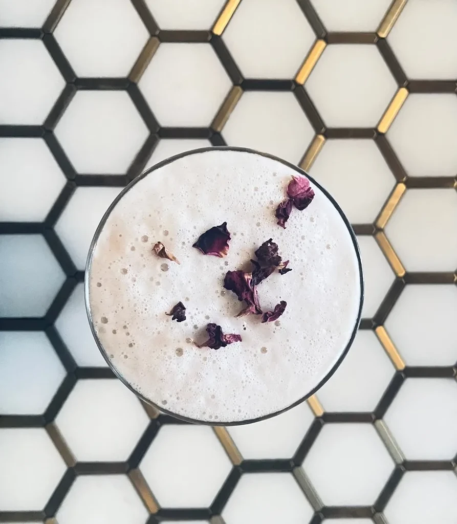 Overhead foamy cocktail with purple petals on hexagonal tiles.