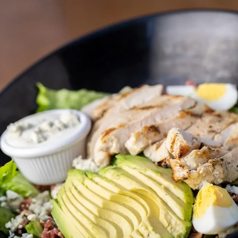 Grilled chicken salad with avocado, eggs, and dressing in a dark bowl.