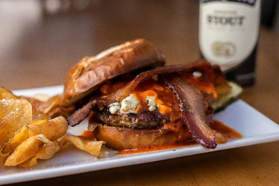 Pretzel bun burger with bacon, blue cheese, hot sauce, and chips.