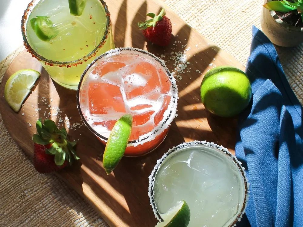 Four colorful, garnished cocktails on a wooden board with fruit.