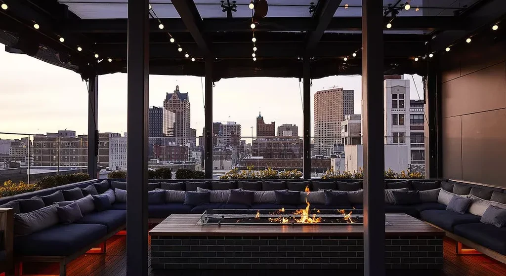 Empty rooftop lounge with fire pit, sofas, string lights, city view.