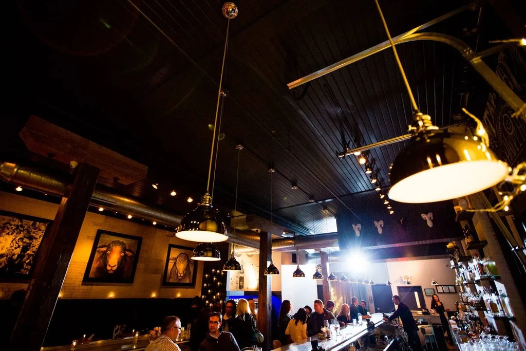 Dimly lit bar interior, industrial-chic ceiling, patrons, and sheep art.