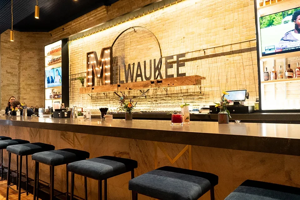 Well-lit modern bar with "MILWAUKEE" sign, cocktails, and bar stools.