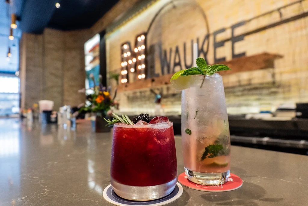 Two vibrant cocktails on a bar counter with a blurred Milwaukee sign.