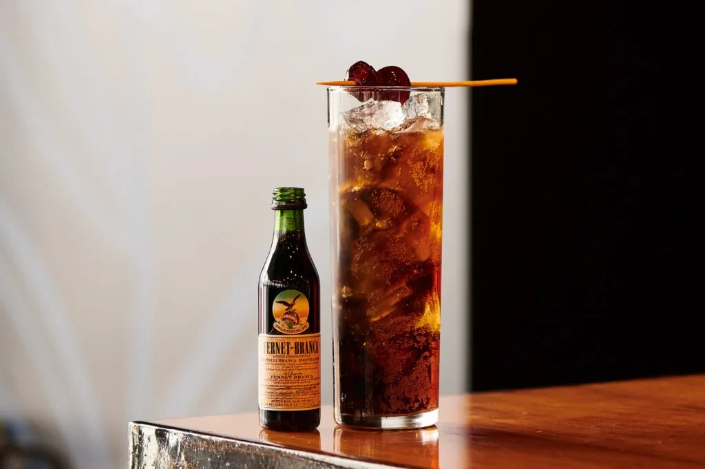 Dark amber cocktail with cherries next to a Fernet-Branca bottle.