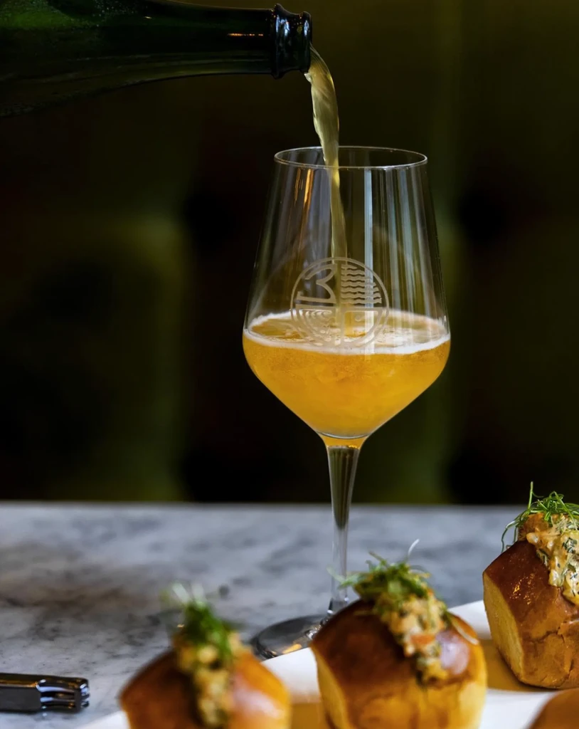 Golden drink poured from green bottle into wine glass with savory items.