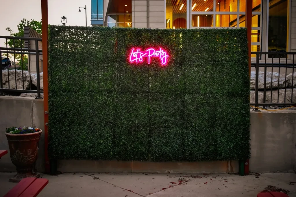Green hedge wall with a pink "Let's Party" neon sign.