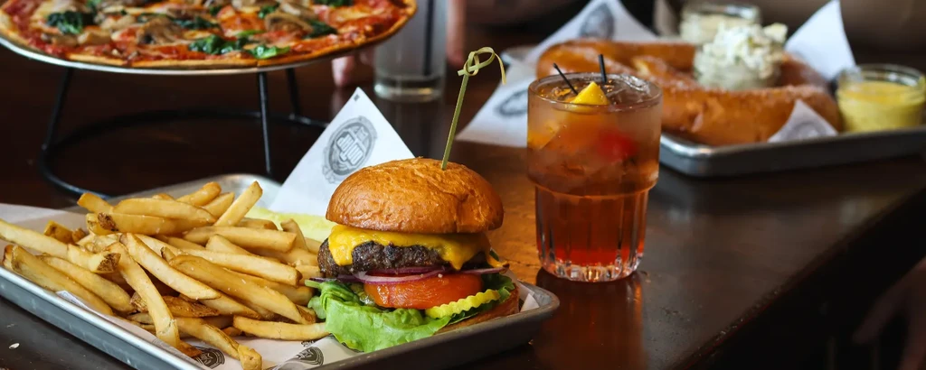 Cheeseburger, fries, cocktail, pizza, and pretzels on a dark restaurant table.