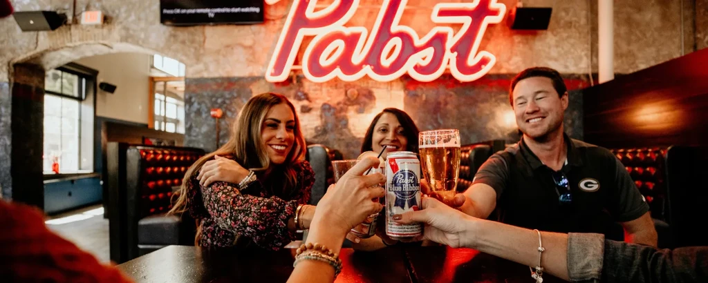 People toasting with drinks, Pabst beer and sign visible in bar.