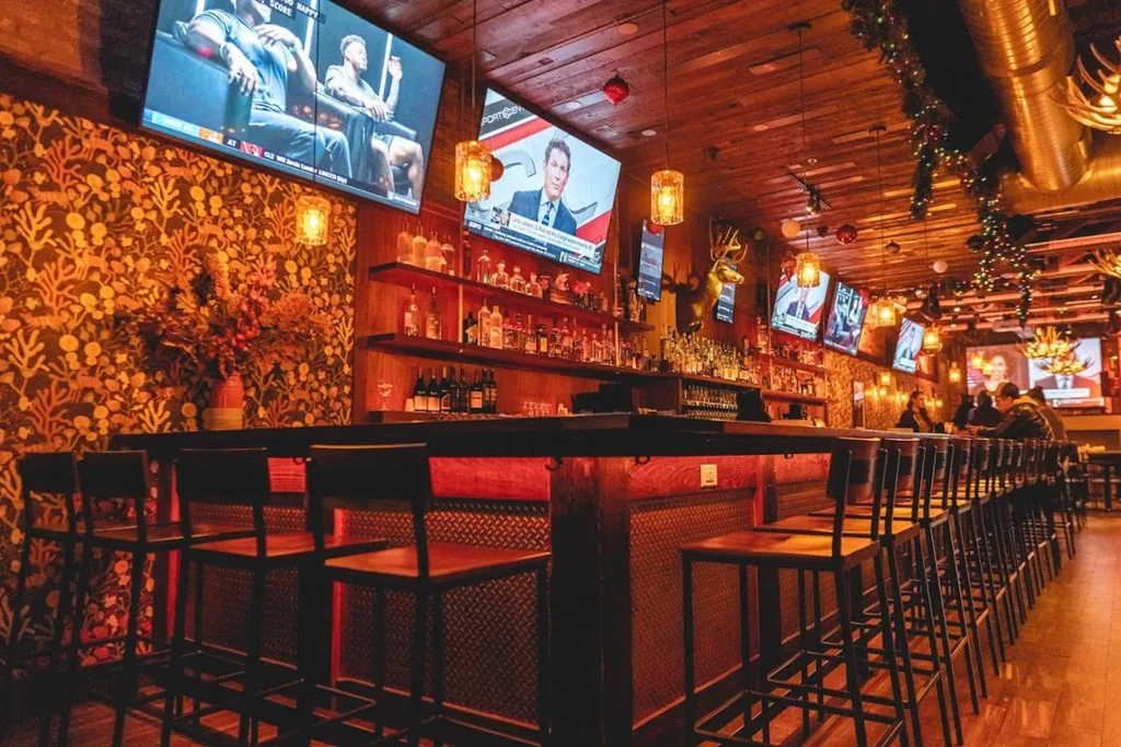 Long dark wood bar counter, stools, liquor, multiple TVs, and garlands.
