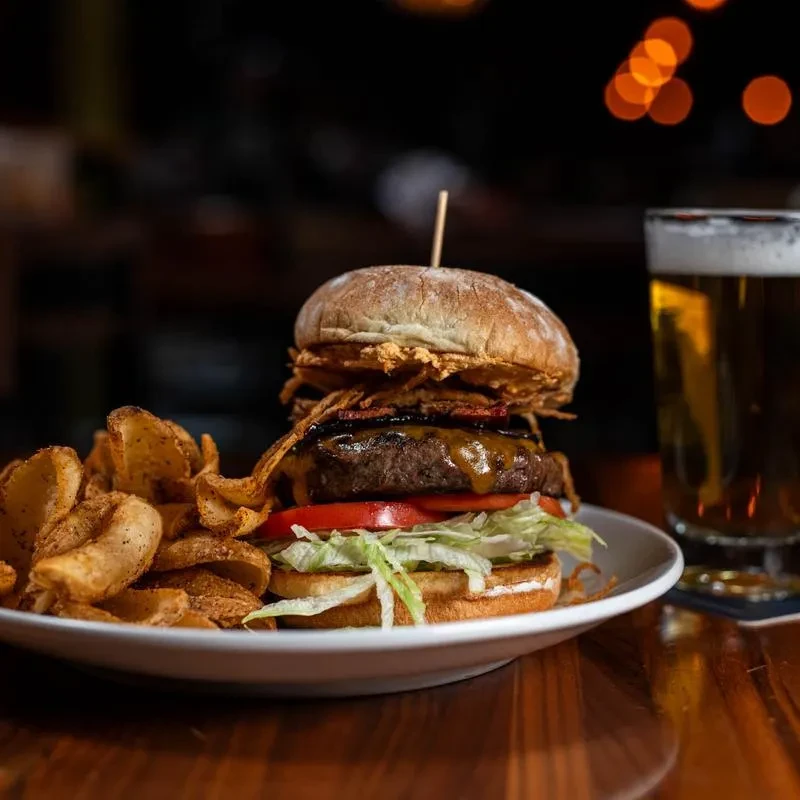 Tall burger, seasoned wedge fries, and amber beer on a wooden table.
