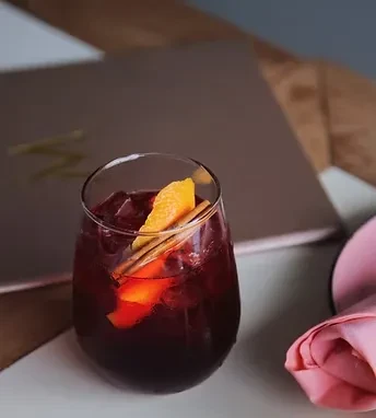 Dark red drink with ice, orange peel, and cinnamon stick.