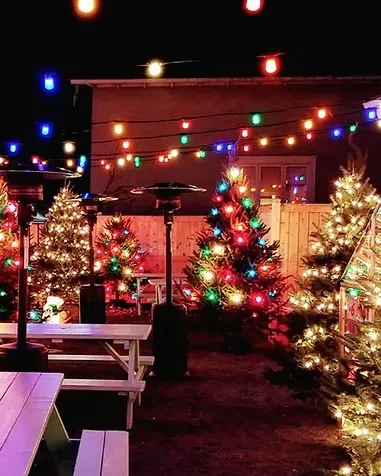 Festive night patio with string lights, Christmas trees, tables, and heaters.