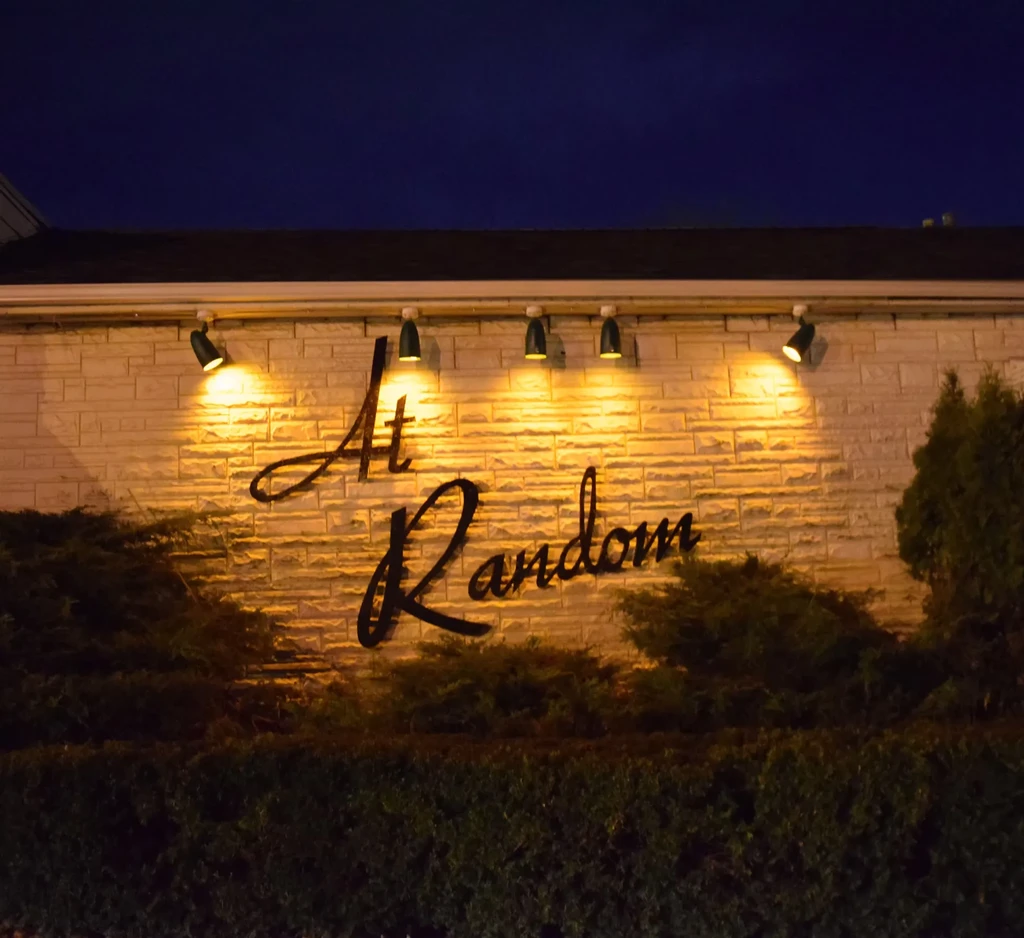 Night photo of 'At Random' sign on brick wall, lit by spotlights.