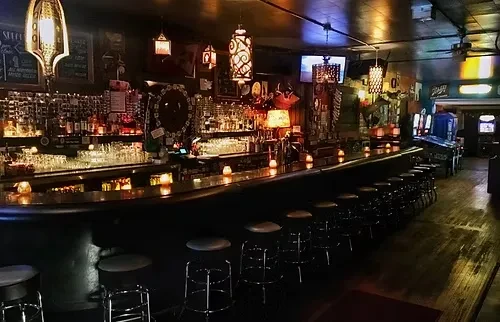Dimly lit, empty bar interior with counter, bottles, and arcade games.