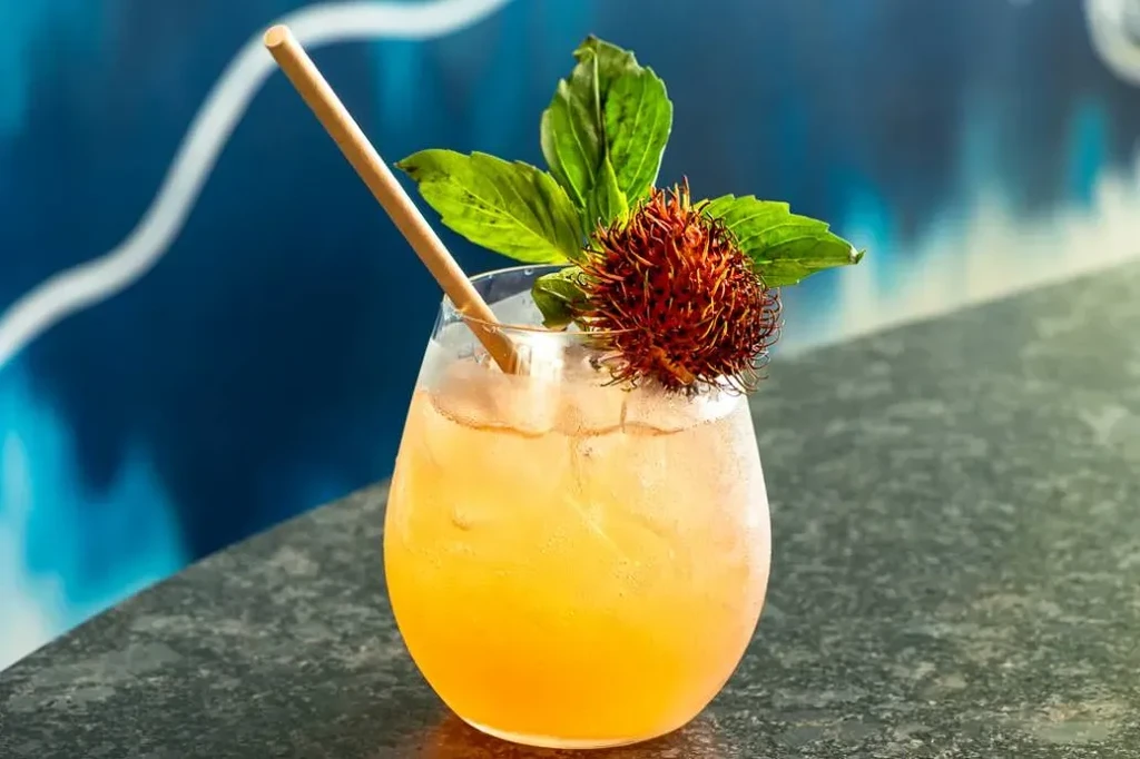 Orange-yellow cocktail with ice, green leaves, and a spiky red fruit.