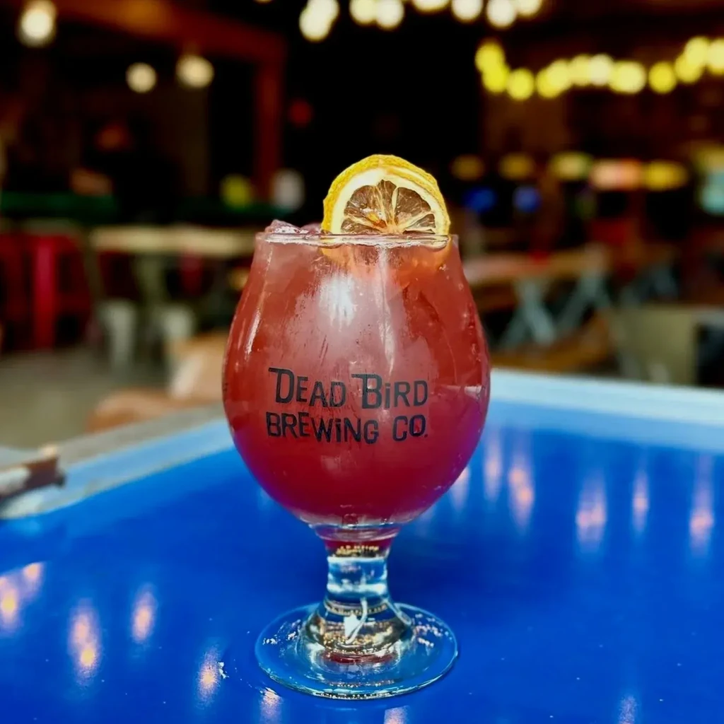 Red-pink cocktail in "DEAD BIRD BREWING CO." glass with dried lemon.