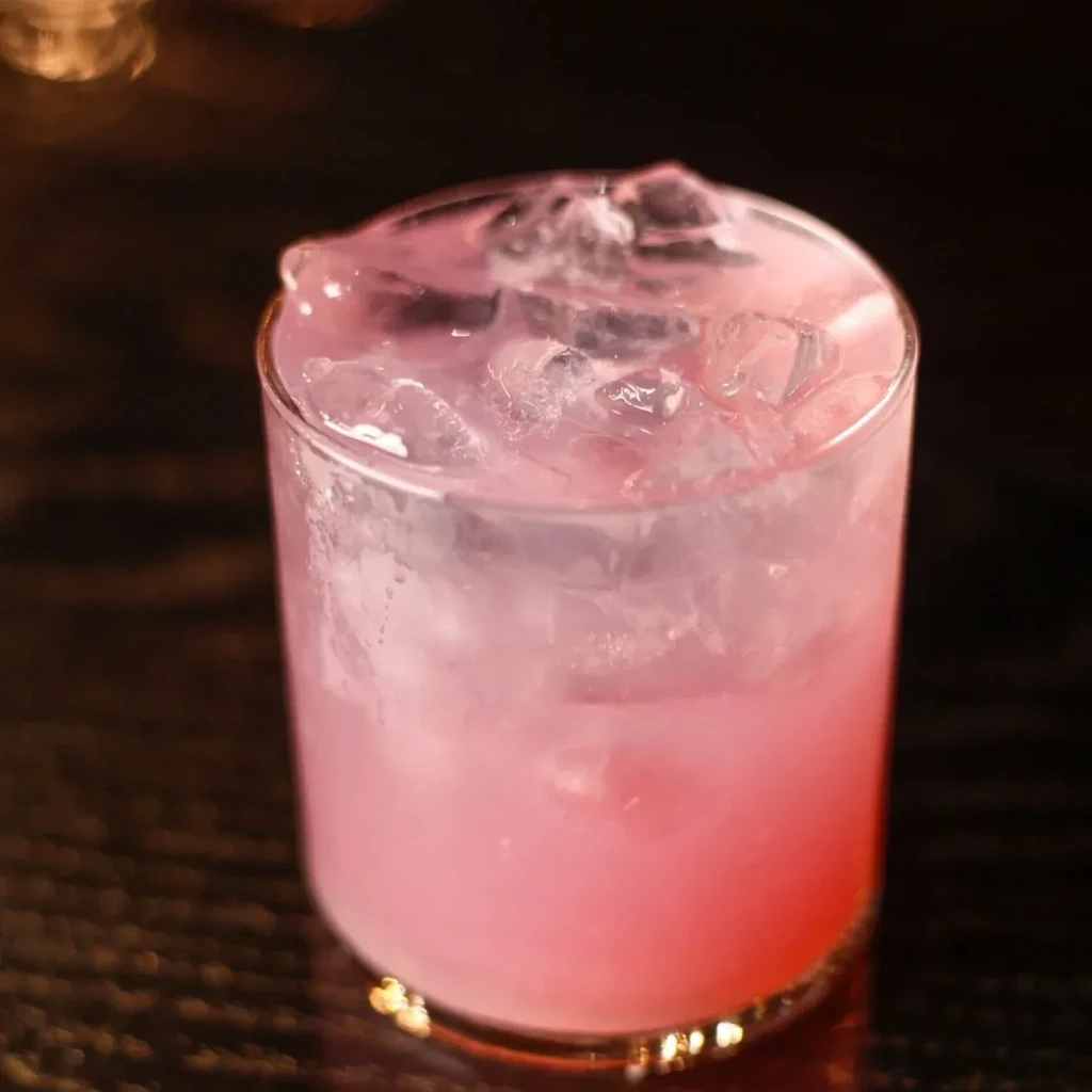 Pink gradient drink with ice in clear glass on dark table.