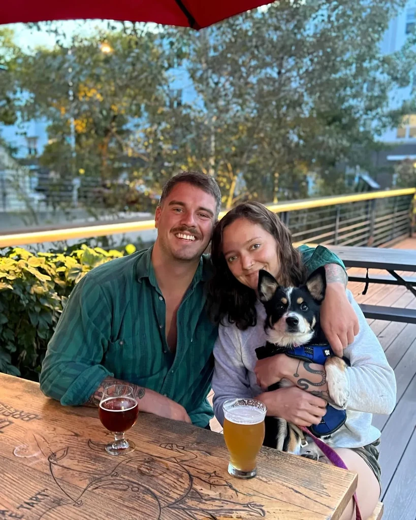 Smiling couple and dog at an outdoor patio table with drinks.